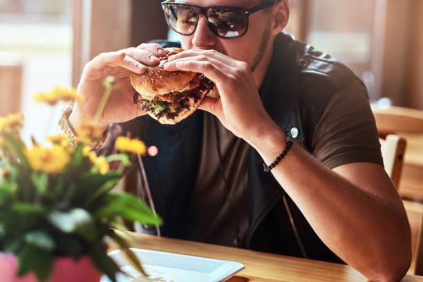 istock Handsome hipster with a stylish haircut and beard sits at a table, decided to dine at a roadside cafe, eating a hamburger. 1028417618