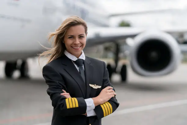 Portrait of a beautiful female pilot next to an airplane and looking at the camera smiling with arms crossed - travel concepts