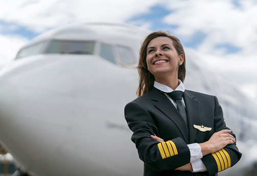 Portrait of a successful female pilot next to an airplane and looking up at the sky smiling with arms crossed - travel concepts