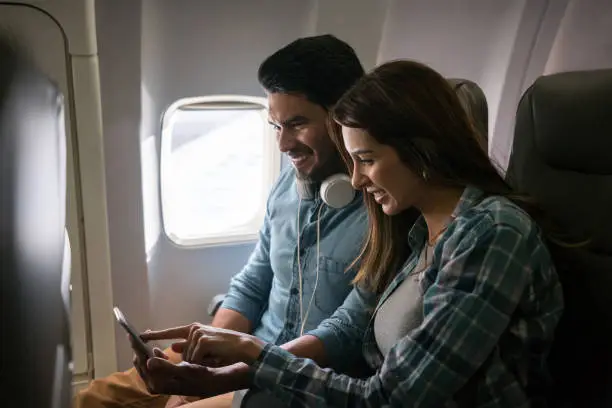 Happy Latin American couple traveling by plane and using their cell phone onboard on airplane mode - lifestyle concepts