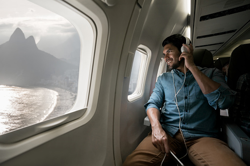 Portrait of a happy man traveling by plane and listening to music on his cell phone with headphones and looking through the window - lifestyle concepts
