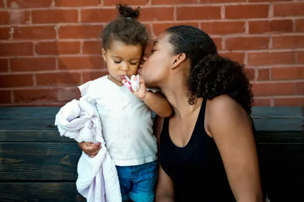 Beautiful mixed-race sisters. One teenager, one toddler. They are relaxed, sitting outdoors in front of a brick wall. They are interacting with each other, big sister kissing baby sister who is holding on her pacifiers and security blanket. Horizontal mid-waist shot with copy space.