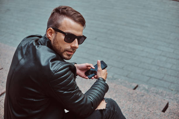 istock Fashionable guy dressed in a black jacket and jeans holds a smartphone sitting on steps. 1028439018