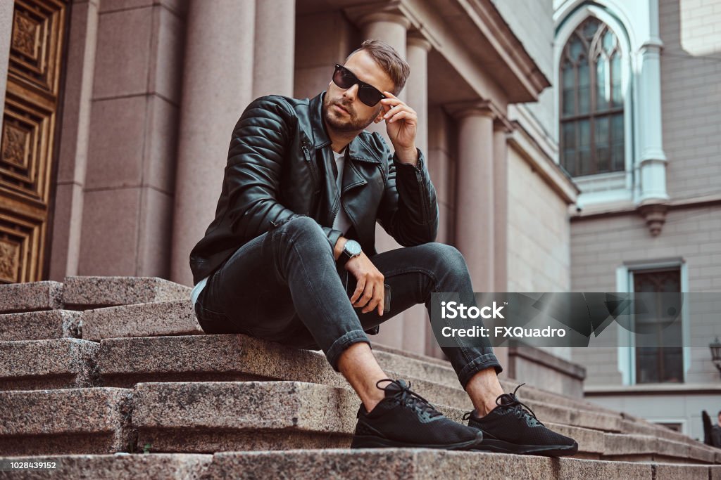 Fashionable guy dressed in a black jacket and jeans holds the smartphone sitting on steps against an old building in Europe. Fashionable guy dressed in a black jacket and jeans holds a smartphone sitting on steps against an old building in Europe. Men Stock Photo