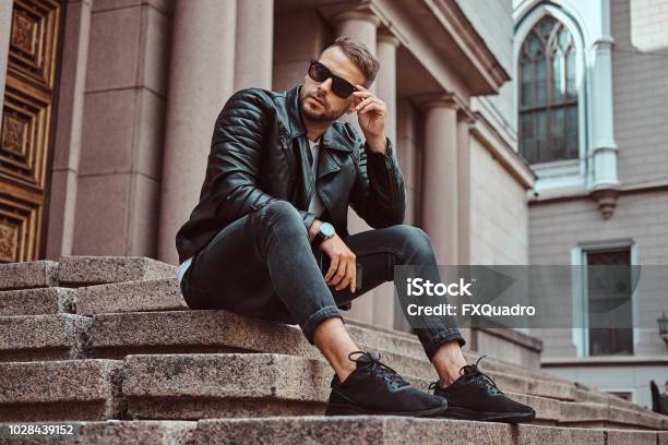 Fashionable Guy Dressed In A Black Jacket And Jeans Holds The Smartphone Sitting On Steps Against An Old Building In Europe Stock Photo - Download Image Now