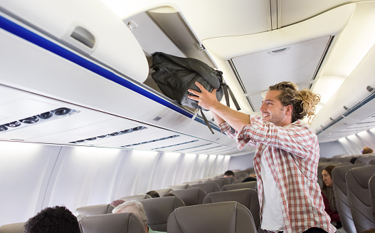 Happy man in an airplane putting his carry-on luggage in the overhead compartment and smiling - travel concepts