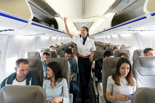 Happy flight attendant walking the aisle in an airplane closing overhead compartments and smiling - travel concepts