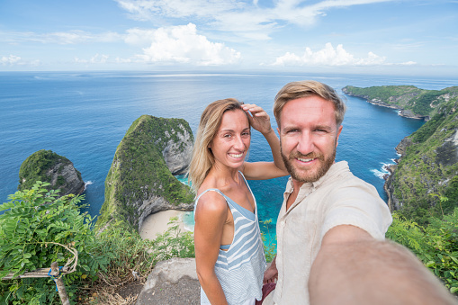 Young couple taking selfie at Kelingking beach from above - Blues and green sand the beautiful mountain shaped on the sea.Travel destinations vacations people love sharing social media concept nature scenics