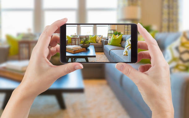 mani femminili che tengono lo smartphone che mostra la foto del soggiorno interno della casa dietro. - screen shoot foto e immagini stock