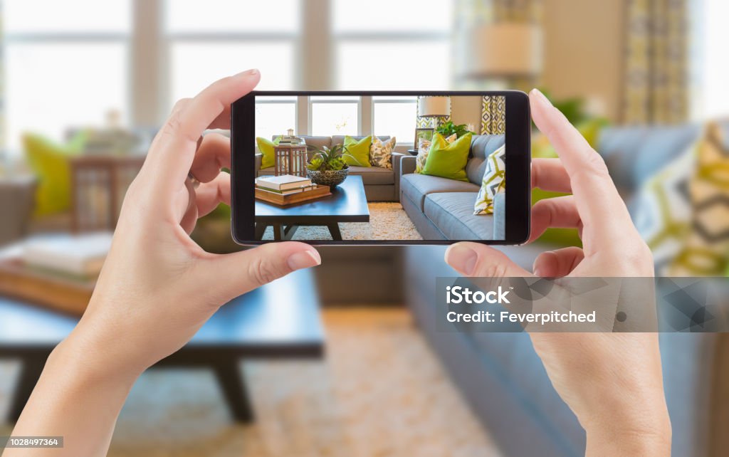 Female Hands Holding Smart Phone Displaying Photo of House Interior Living Room Behind. Photographing Stock Photo