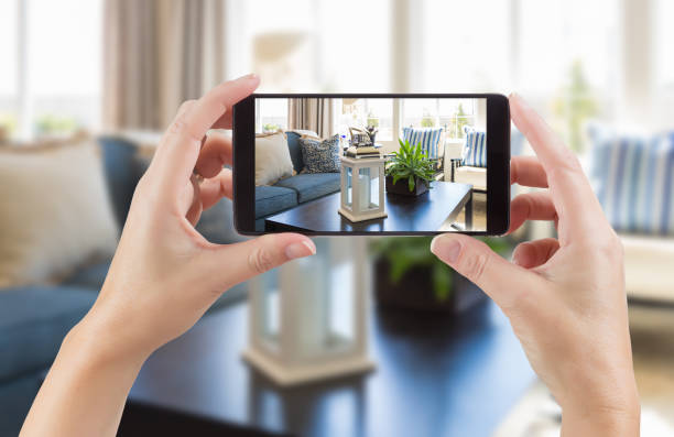 Female Hands Holding Smart Phone Displaying Photo of House Interior Living Room Behind. stock photo