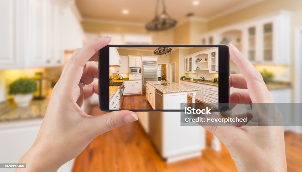 Female Hands Holding Smart Phone Displaying Photo of Kitchen Behind. Photographing Stock Photo