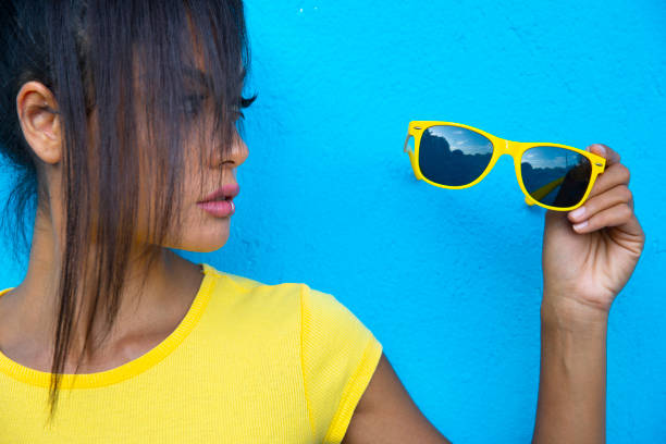Asian woman holding sunglasses in front of colored background.
