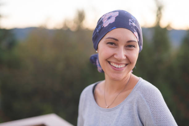 Portrait of a beautiful woman with cancer A beautiful young woman wearing a head wrap looks toward the camera and smiles radiantly. She is standing outdoors and there are mountains and trees in the background. survival stock pictures, royalty-free photos & images