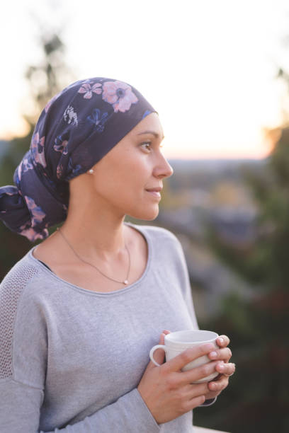 woman battling cancer stands outside and contemplates her life A beautiful young woman wearing a head wrap looks off in the distance and smiles contemplatively while drinking a cup of tea. She is standing outdoors and there are mountains and trees in the background. cancer illness stock pictures, royalty-free photos & images