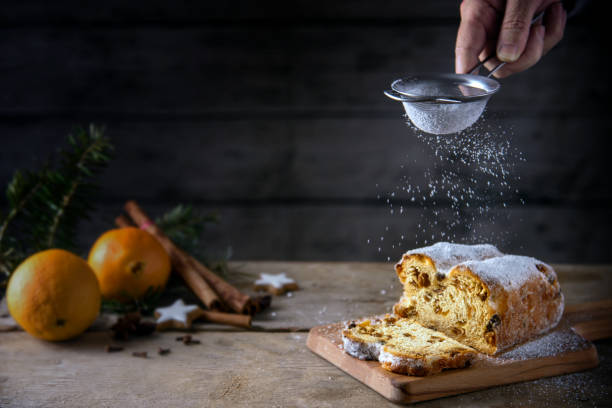 Sieving powdered sugar on a christmas cake, in germany christstollen, orange and spices blurred in the back on a rustic wooden table, dark background with copy space Sieving powdered sugar on a christmas cake, in germany christstollen, orange and spices blurred in the back on a rustic wooden table, dark background with copy space, selected focus, narrow depth of field sprinkling powdered sugar stock pictures, royalty-free photos & images
