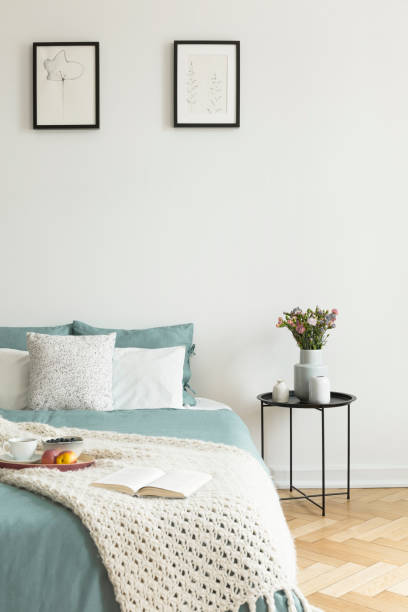 istock Pale sage and white linen, pillows and a blanket on a bed standing against white wall with two pictures in a bedroom interior. Black metal side table by the bed. Real photo 1028739780