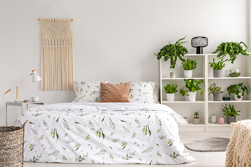 Close to nature bright bedroom interior with many green plants beside a big bed. Woven tapestry above the bed. Real photo.
