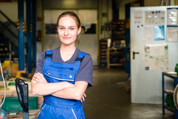 retrato de sonriente mujer joven aprendiz en industria del metal, brazos cruzados - mono ropa protectora fotografías e imágenes de stock