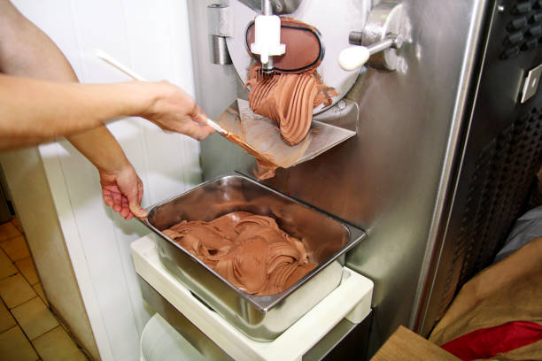 istock Confectioner in chef uniform is working on ice cream maker machine. Producing black chocolate ice cream flavors and it falls into steel container. Industrial preparation of italian creamy ice cream. 1028806452