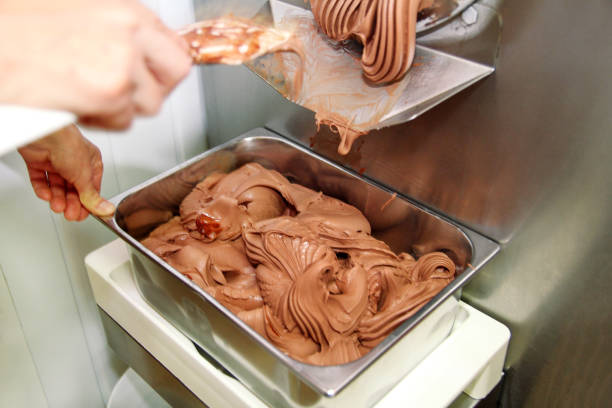 istock Confectioner in chef uniform is working on ice cream maker machine. Producing black chocolate ice cream flavors and it falls into steel container. Industrial preparation of italian creamy ice cream. 1028806466