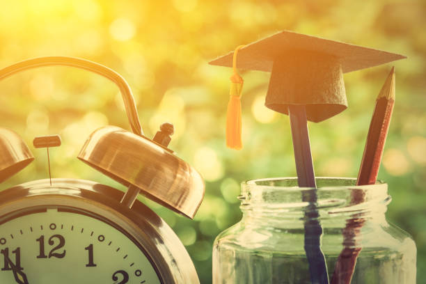 Education is the most powerful weapon or knowledge is power concept : Black graduation cap or hat with tassel hangs on a pencil in a clear glass jar or a bottle with red and blue pencils near a clock.