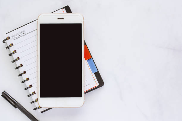 istock Smartphone with spiral notebook and black pen on marble background for media technology and workspace concept 1028837350