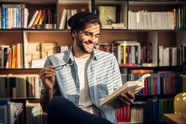 istock Cute smiling hipster boy sitting on a library floor and looking through some books. 1028869474