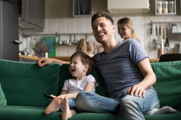 Happy dad and kid son laughing watching tv together Happy dad and kid son holding remote control laughing at funny humor comedy film or tv show sitting on sofa at home, smiling father having fun with child boy watching television together on weekend part of a series photos stock pictures, royalty-free photos & images