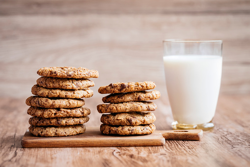 A glass of milk and some homemade cookies, with melted chocolate chips. The cookies are piled up on a on a little wooden cutting board, lying on a wooden table. The background is wooden as well.