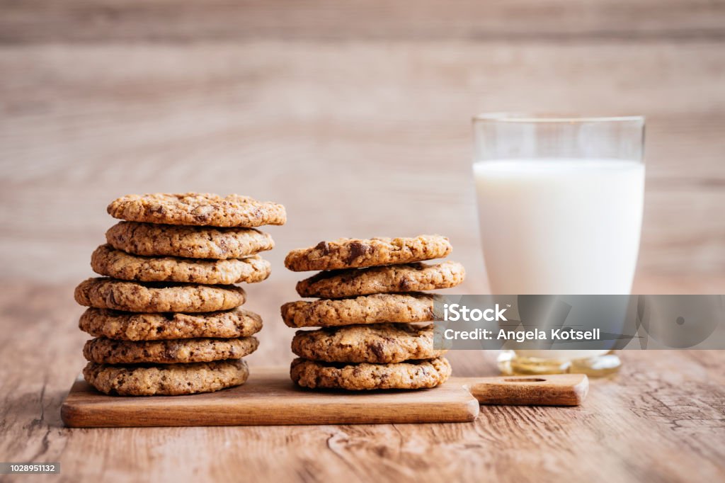 Milk and cookies, homemade with chocolate chips. A glass of milk and some homemade cookies, with melted chocolate chips. The cookies are piled up on a on a little wooden cutting board, lying on a wooden table. The background is wooden as well. Cookie Stock Photo