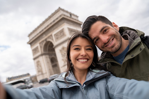 Portrait of a happy couple sightseeing in Paris and taking a selfie by the Arc de Triomphe while smiling - people traveling concepts