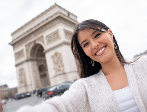 Portrait of a happy female tourist in Paris taking a selfie with the Arc de Triomphe and smiling - people traveling concepts