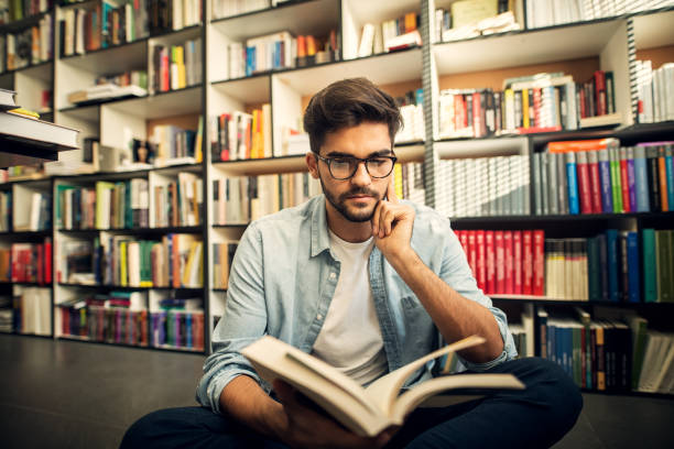 istock Cute smiling hipster boy sitting on a library floor and looking through some books. 1029194704