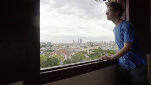 istock Young Man Looking Out Window With City in Background 1029252342