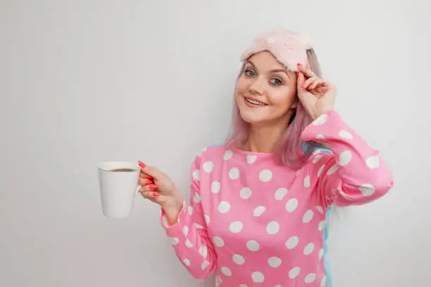 Happy young woman drink morning coffee. Beautiful girl in pink pajamas and mask for sleep