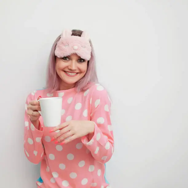 Happy young woman drink morning coffee. Beautiful girl in pink pajamas and mask for sleep