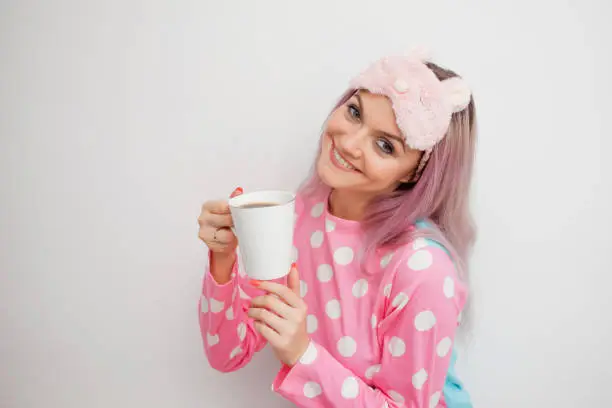 Happy young woman drink morning coffee. Beautiful girl in pink pajamas and mask for sleep