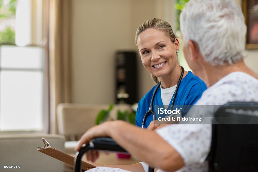 Friendly nurse talking to senior patient Friendly doctor examining health of patient sitting in wheelchair. Happy smiling nurse consulting disabled patient about treatment. Nurse caring about elder handicap man at home. Nurse Stock Photo