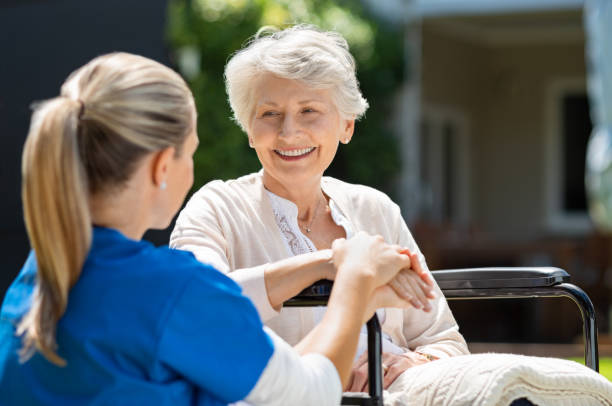 Nurse takes care of old patient Smiling senior patient sitting on wheelchair with nurse supporting her. Doctor looking at elderly patient on a wheelchair in the garden. Nurse holding hand of mature woman outside pension home. persons with disabilities photos stock pictures, royalty-free photos & images