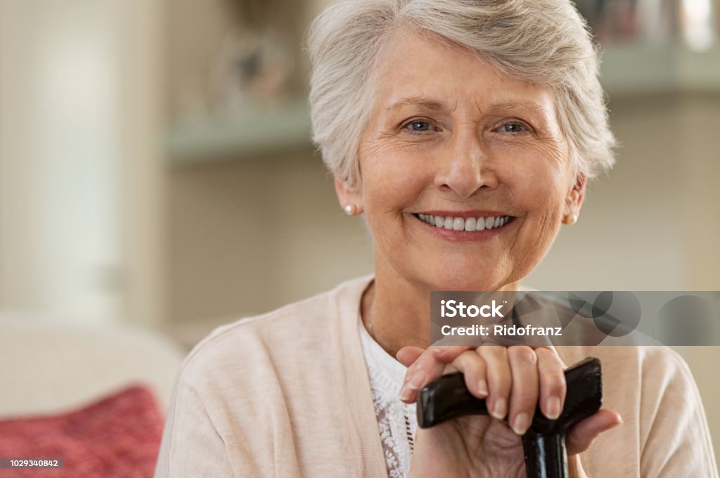 Elderly woman smiling at home Retired woman with her wooden walking stick at home. Happy senior woman relaxing at home holding cane and looking at camera. Smiling grandmother sitting on couch. Senior Adult Stock Photo