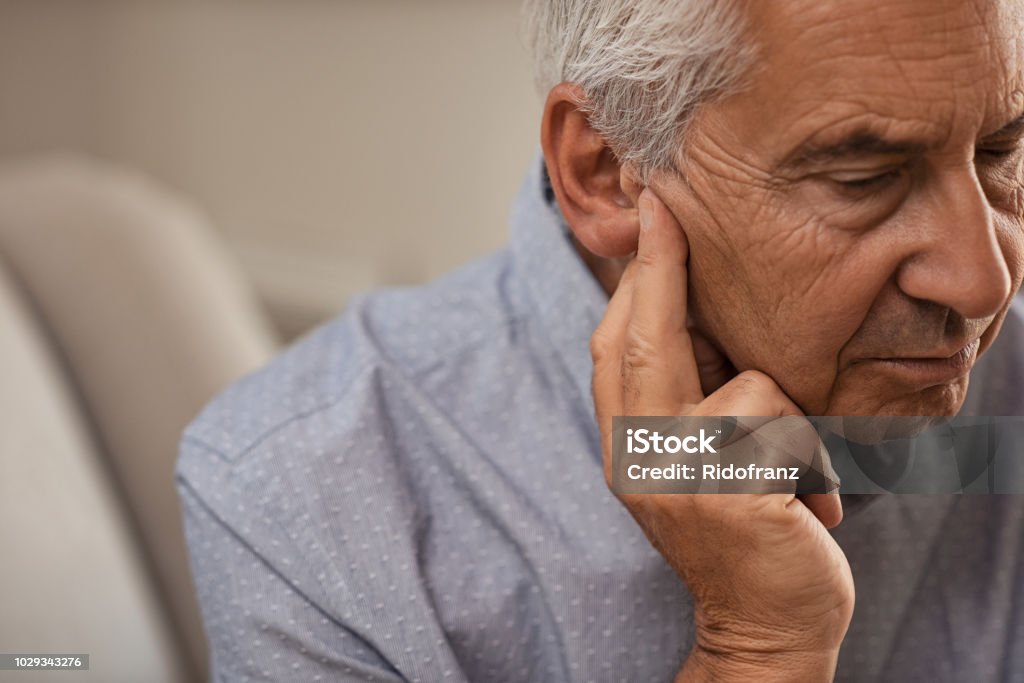 Senior man with hearing problems Side view of senior man with symptom of hearing loss. Mature man sitting on couch with fingers near ear suffering pain. Listening Stock Photo