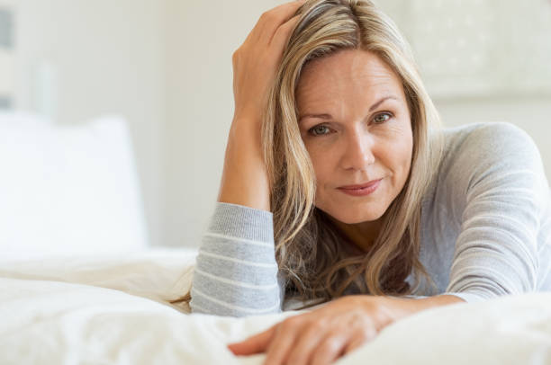 Thoughtful woman laying on bed in bedroom and looking at camera. Portrait of middle aged woman smiling. Portrait of a beautiful middle aged woman at home.