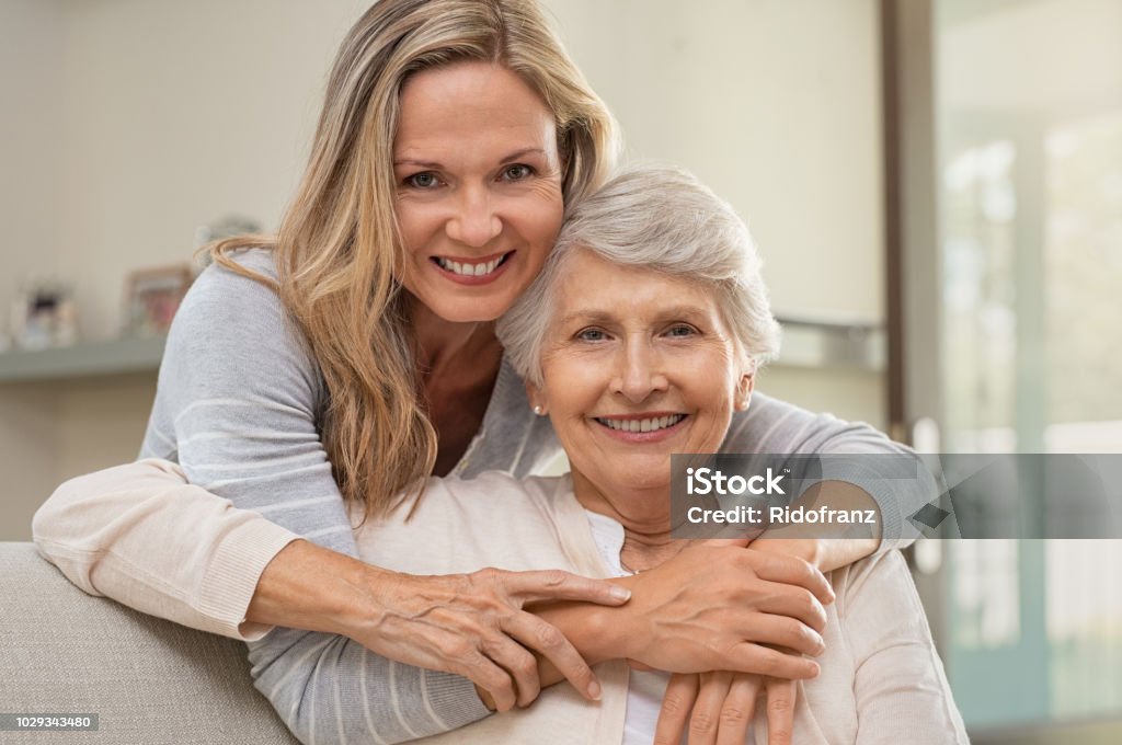 Woman hugging mother with love Cheerful mature woman embracing senior mother at home and looking at camera. Portrait of elderly mother and middle aged daughter smiling together. Happy daughter embracing from behind elderly mom sitting on sofa. Daughter Stock Photo