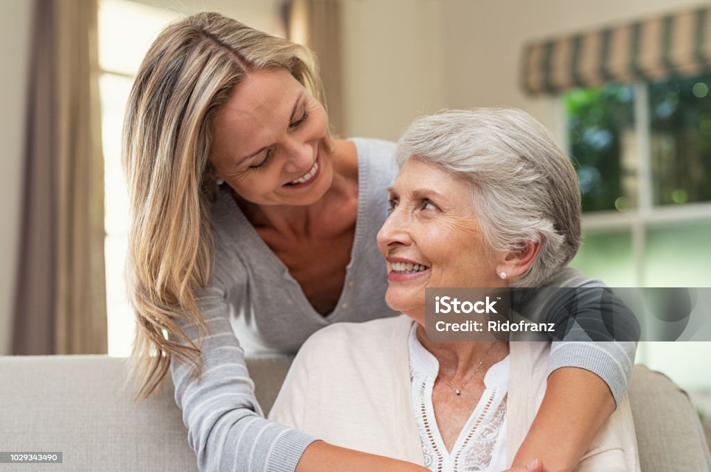 Woman embracing senior mother Portrait of old mother and mature daughter hugging at home. Happy senior mother and adult daughter embracing with love on sofa. Cheerful woman hugging from behind older mom and looking at each other. Senior Adult Stock Photo