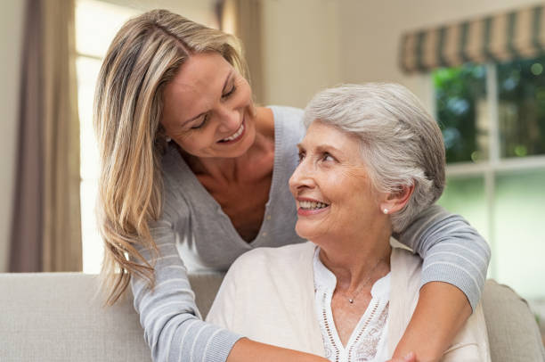 Woman embracing senior mother Portrait of old mother and mature daughter hugging at home. Happy senior mother and adult daughter embracing with love on sofa. Cheerful woman hugging from behind older mom and looking at each other. arm around photos stock pictures, royalty-free photos & images