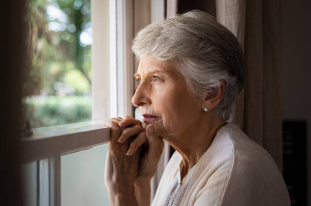 Lonely senior woman Depressed senior woman at home feeling sad. Elderly woman looks sadly outside the window. Depressed lonely lady standing alone and looking through the window. looking through window stock pictures, royalty-free photos & images