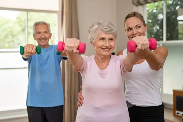 Happy senior couple exercising with dumbbells. Mature personal trainer helping elderly woman exercise at home. Old man and woman doing stretching exercise with physiotherapist.