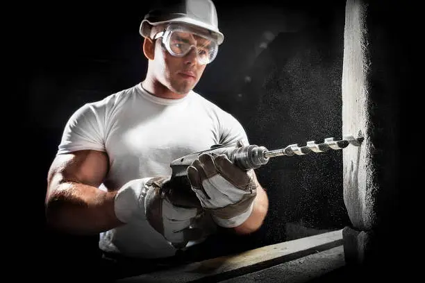 Man with a tool. Worker using a drilling power tool on construction site. Young man using electric drill on white brick wall in a white helmet.