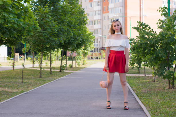 Full-length portrait of fashionably dressed young female standing on city street. Stunning girl in red skirt posing outside surrounded with trees. Modern fashion concept